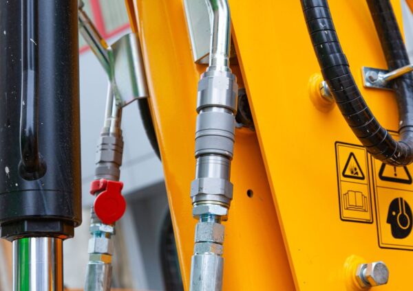 A close-up of hydraulic hoses and metal fittings on heavy machinery with a hydraulic cylinder and safety labels visible.