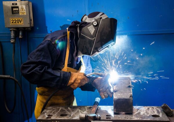 A welder in protective gear uses a torch on a metal component as bright sparks fly in a fabrication workshop.